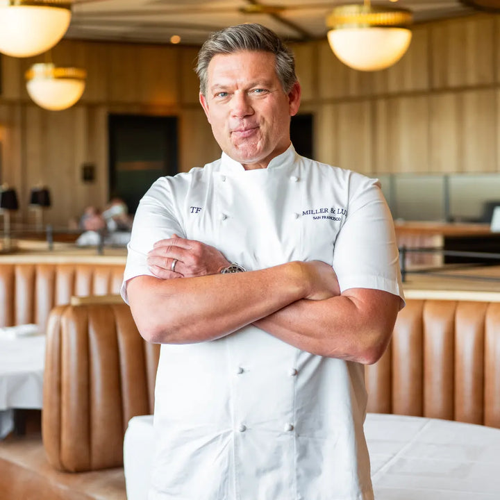 A chef in a white uniform stands with arms crossed in a modern restaurant, featuring tan booths and pendant lighting, with the NanoBond Titanium x Tyler Florence Cookware Set, 41-Piece, gleaming behind him.