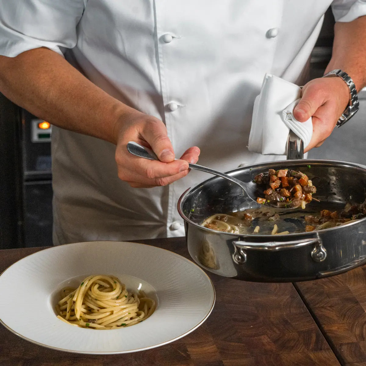 A chef in a white coat serves pasta with sauce from the NanoBond Titanium x Tyler Florence Cookware Set, 41-Piece, into a white bowl with some spaghetti already plated.