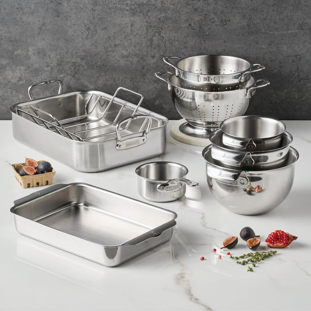 The Hestan Provisions Host and Roast 8-piece set, featuring stainless steel mixing bowls, colander, roasting and baking pans, and a measuring cup, is displayed neatly on a white counter with figs and herbs nearby.