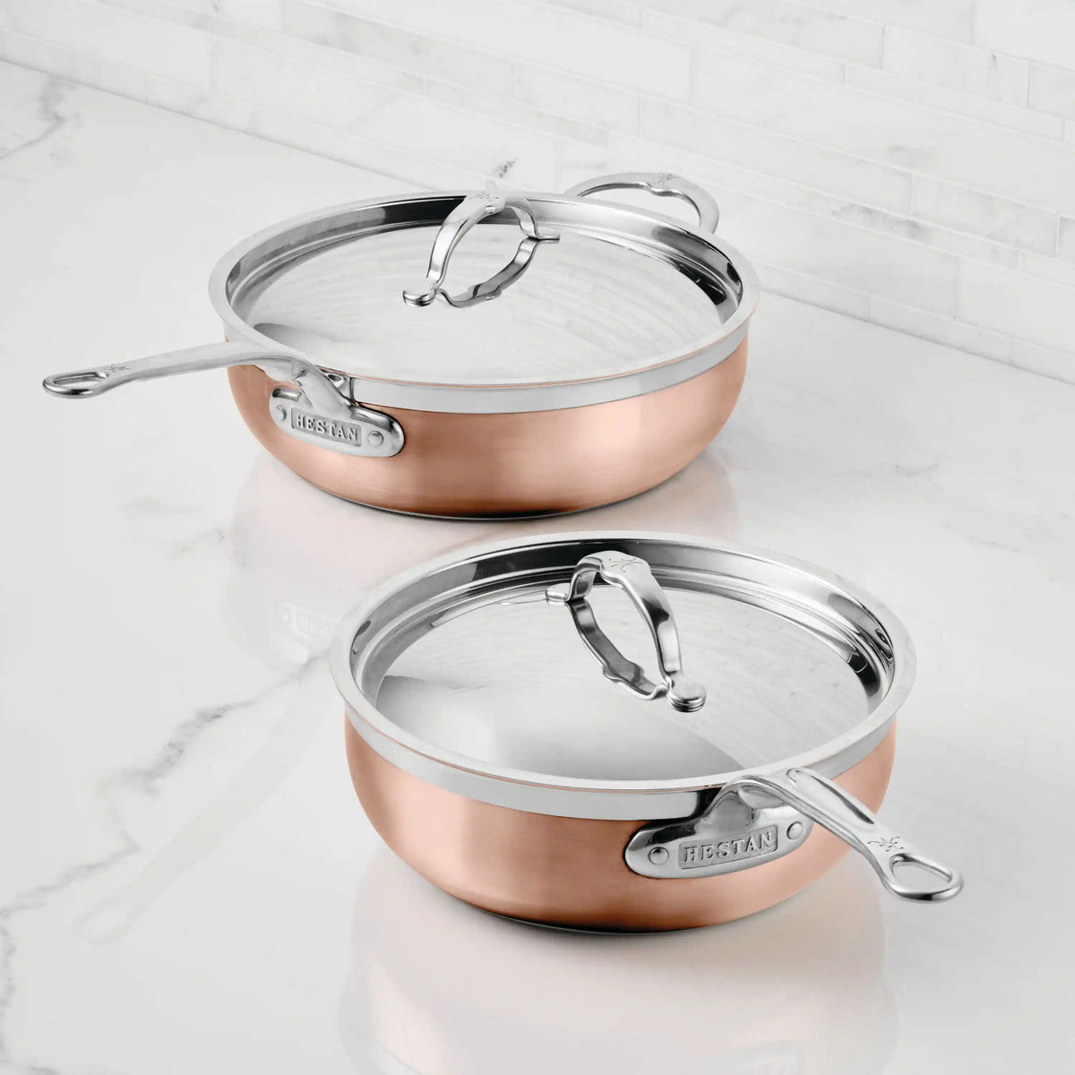 Two CopperBond Copper Induction Essential Pans with lids are on a white marble countertop in front of a white tiled backsplash. The CopperBond brand name is visible on the handles, highlighting their induction compatible design.