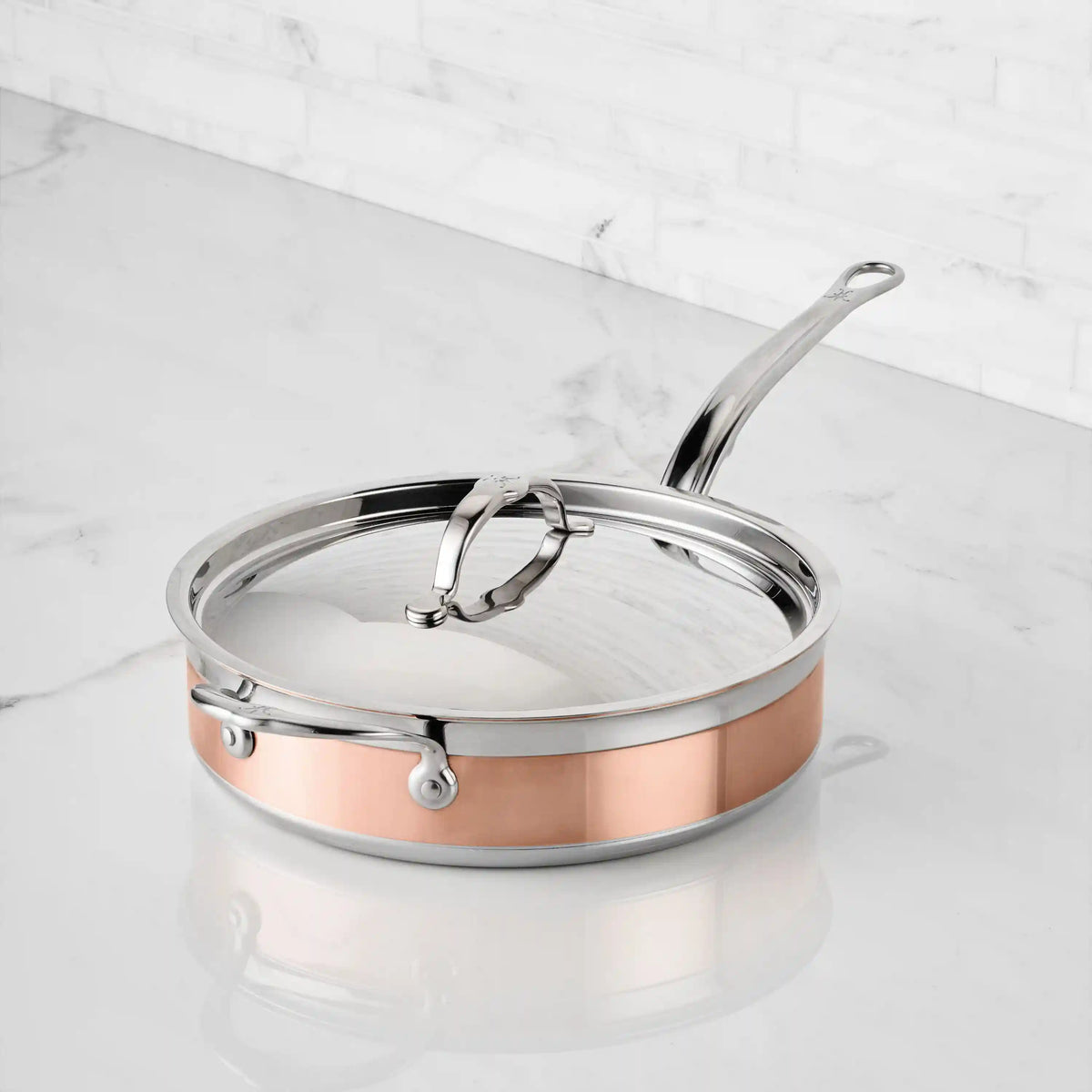 A CopperBond Copper Induction Sauté with Lid, 3.5-Quart, sits on a white marble countertop. It features a polished metal handle, shiny copper band, and durable 18/10 stainless steel construction.