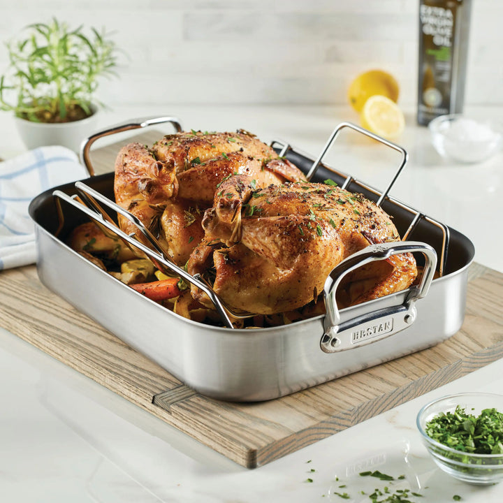 The Hestan Provisions Nonstick Clad Stainless Steel Roasters with Rack holds two seasoned chickens above roasted vegetables, with fresh herbs, lemon, olive oil, and chopped parsley completing this ideal stainless steel cookware setup.