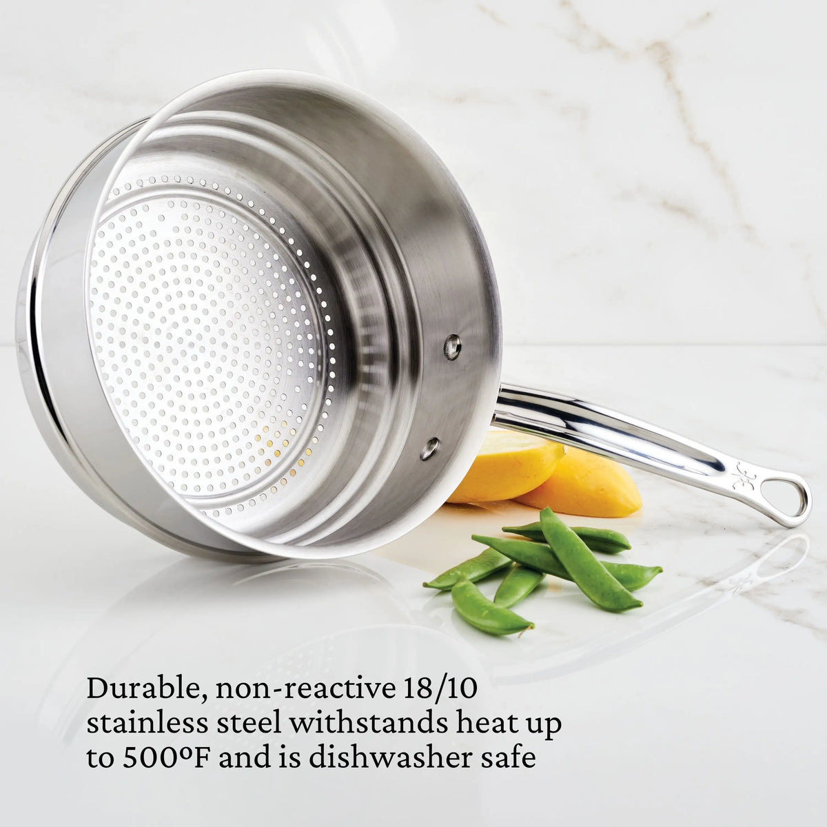 A Thomas Keller Commercial Clad Stainless Steel 29-Piece Cookware Set saucepan sits on white marble with lemon slices and snap peas, featuring highlighted durability, heat resistance, and dishwasher safety.