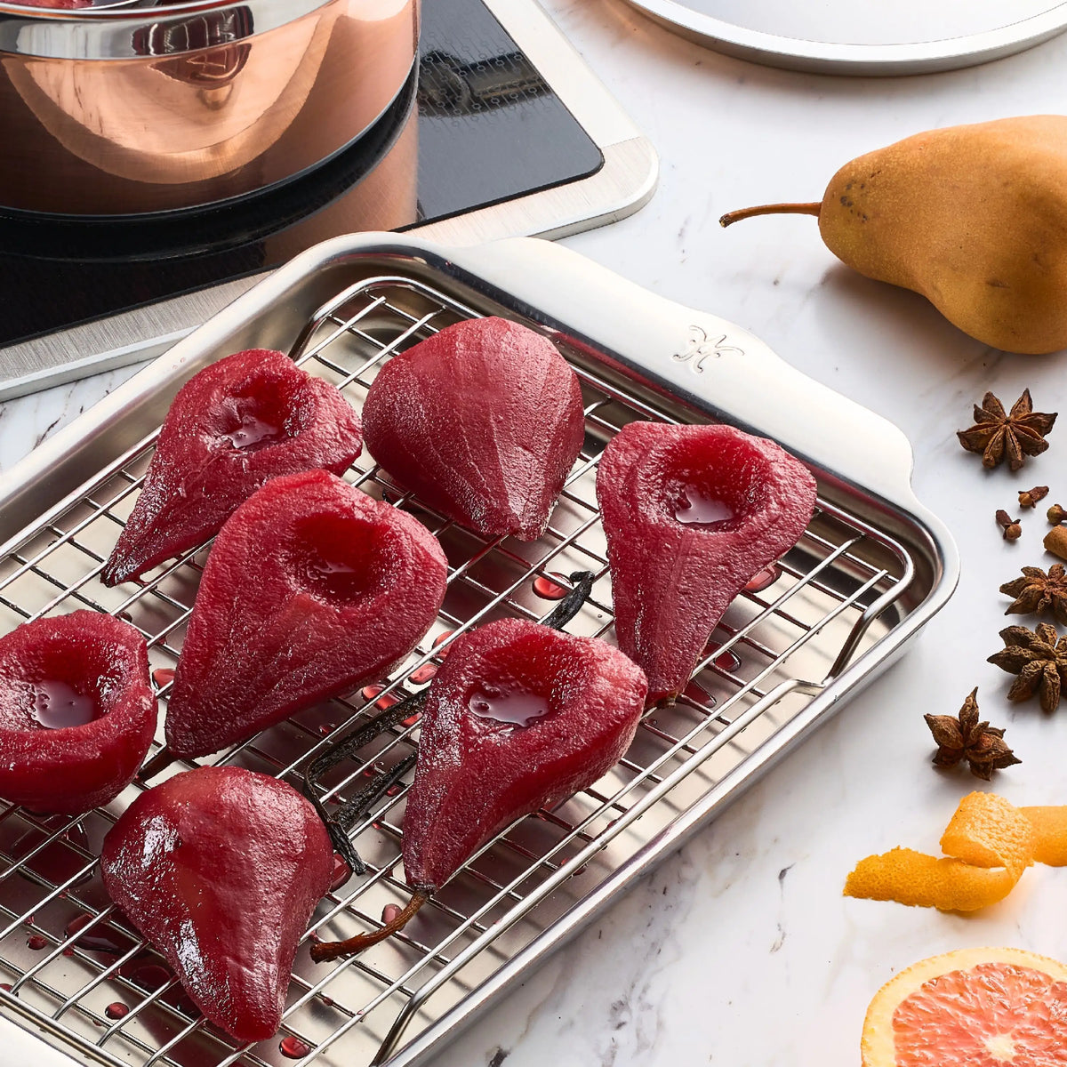 Six halved, poached red pears cool on a rack above a tray. Nearby are a raw pear, orange peel, grapefruit slice, and star anise on marble. In the background is a Thomas Keller Commercial Clad 29-Piece Stainless Steel cookware pot on an induction cooktop.
