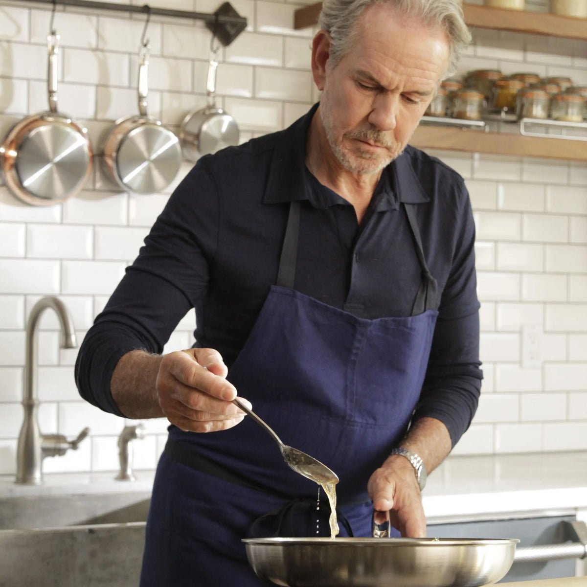 An older man in a navy apron stands in a modern kitchen, drizzling liquid into a metal bowl. Behind him, copper pots and Thomas Keller Commercial Clad Stainless Steel 9-Piece Cookware Set hang on the wall, with spice jars on a shelf.