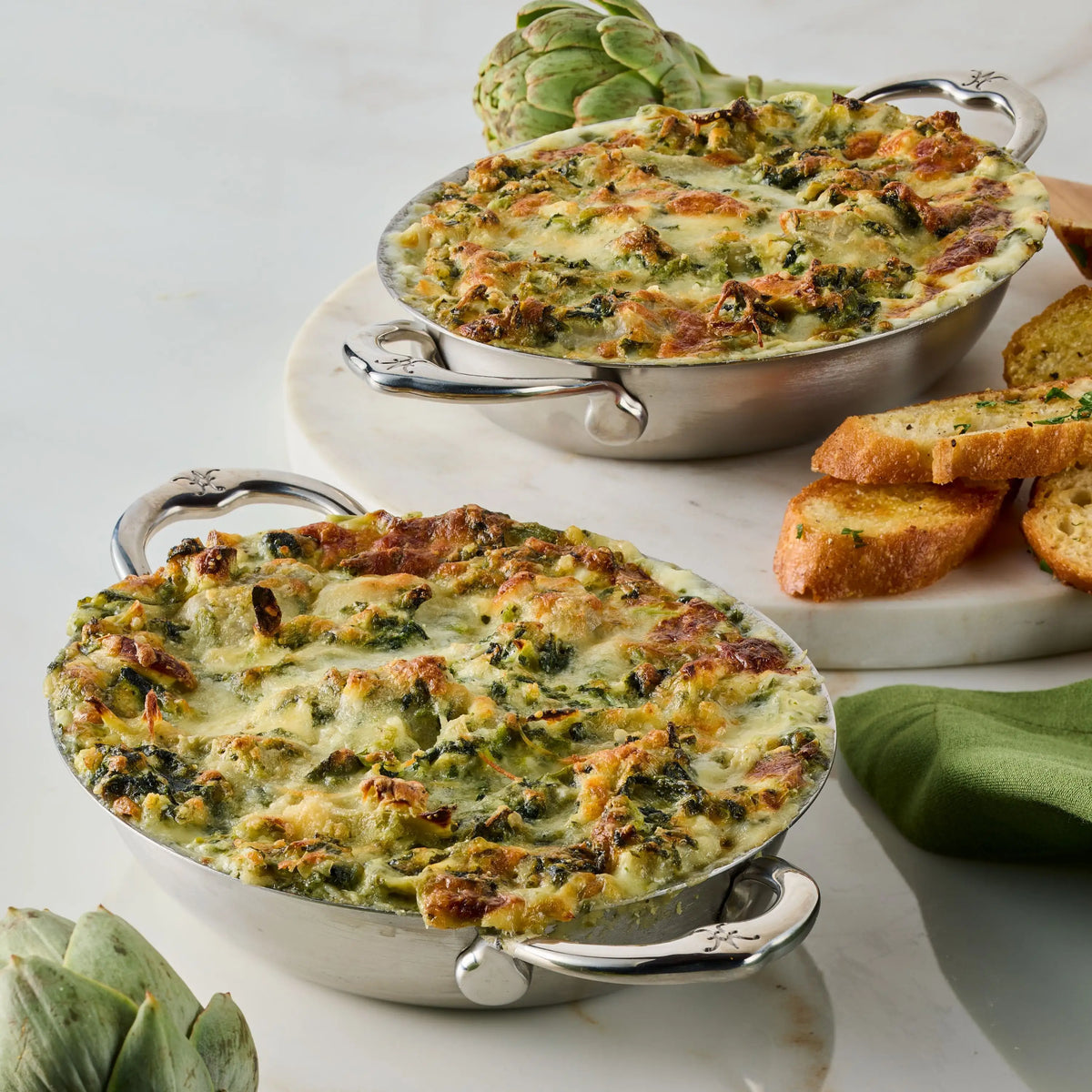 Two oval silver dishes from the Thomas Keller Commercial Clad Stainless Steel 29-Piece Cookware Set are filled with baked spinach and artichoke dip, topped golden brown, set on a white surface with toasted bread, whole artichokes, and a green napkin.