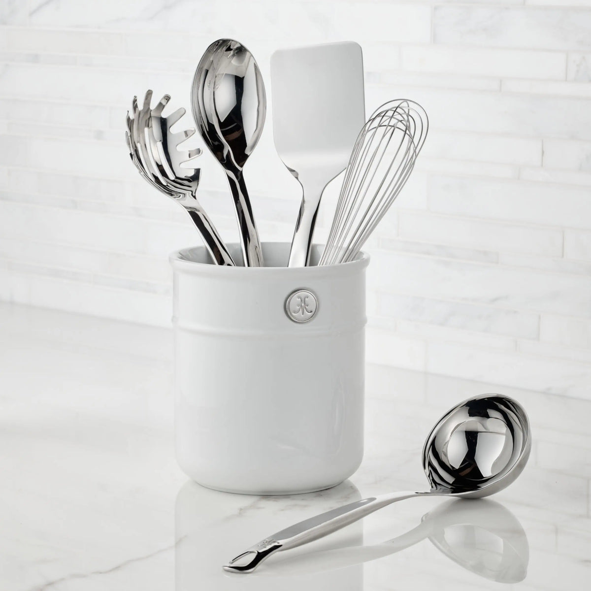A white ceramic utensil holder with five stainless steel tools—spaghetti server, slotted spoon, solid spoon, spatula, and whisk—sits on a marble countertop. A Thomas Keller Commercial Clad ladle from the 29-piece cookware set rests beside it.