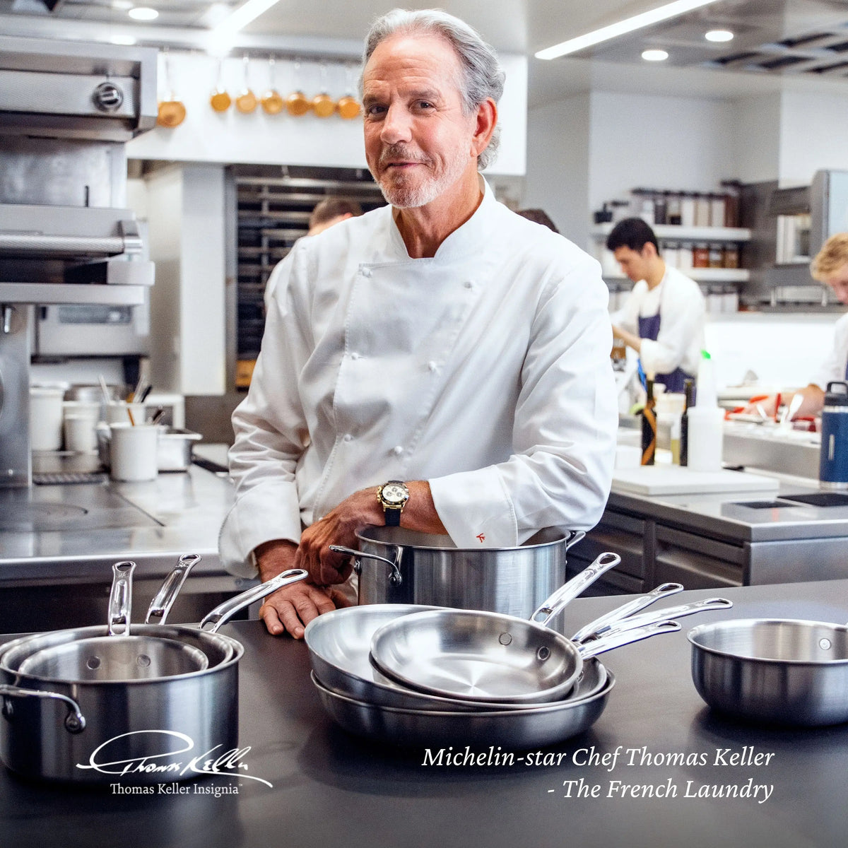 A chef in a white uniform stands in a modern kitchen behind the Thomas Keller Commercial Clad 29-Piece Stainless Steel Cookware Set. Other staff work in the background. Text on the image reads Michelin-star Chef Thomas Keller – The French Laundry.