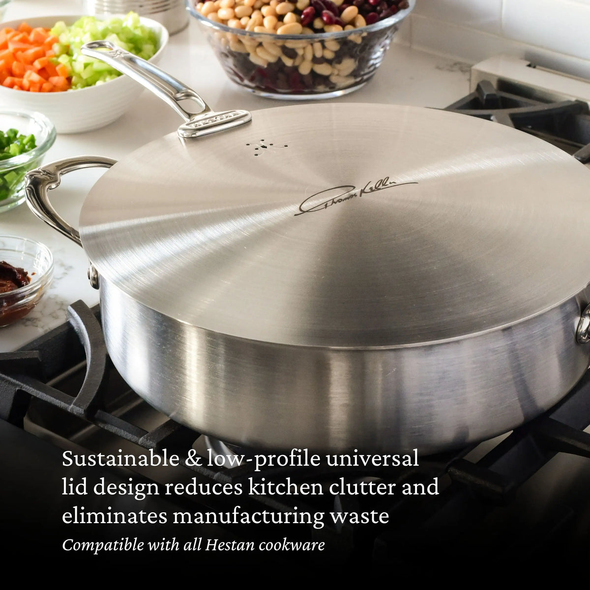 A pan from the Thomas Keller Commercial Clad Stainless Steel 29-Piece Cookware Set with a universal lid is on the stove. Glass bowls with chopped vegetables and beans sit nearby. Text highlights the lid’s sustainability and Hestan compatibility.