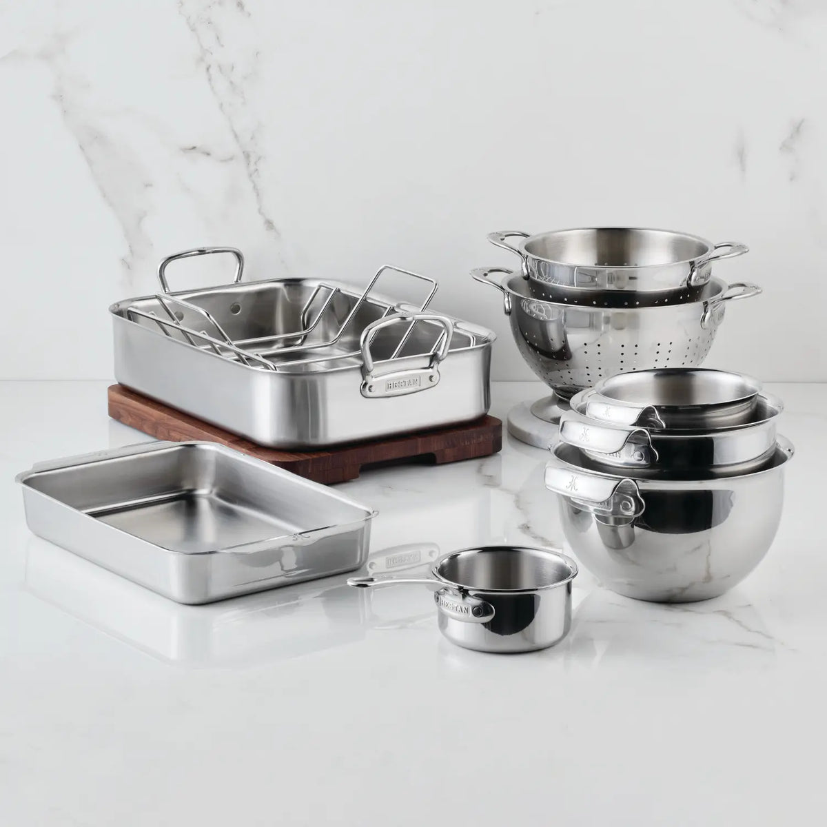 The Hestan Provisions Host and Roast 8-piece Set, featuring stainless steel cookware, mixing bowls, colanders, saucepans, a roasting pan, bakeware, and a wooden cutting board, is displayed on a white marble countertop.