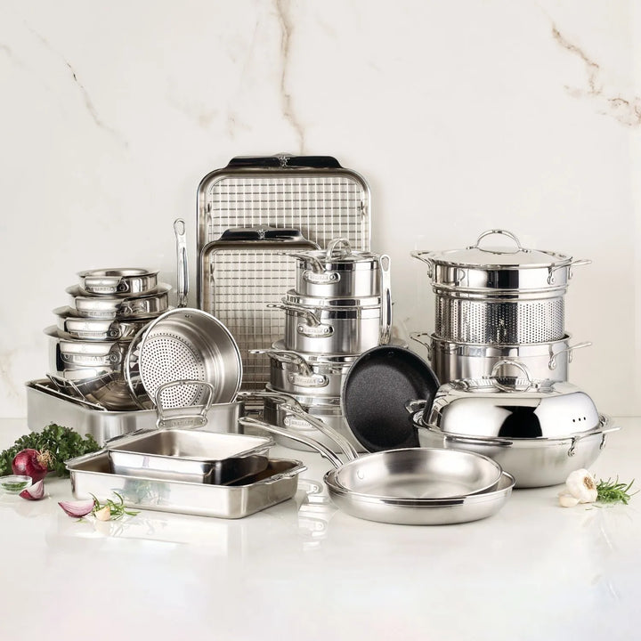 A neatly arranged ProBond Professional Clad Performance 28-piece set, featuring stainless steel cookware with a tri-ply aluminum core, is displayed against a light marble background. The set includes pots, pans, lids, a baking tray, roasting pan, and steamer with onions and garlic scattered in the foreground.