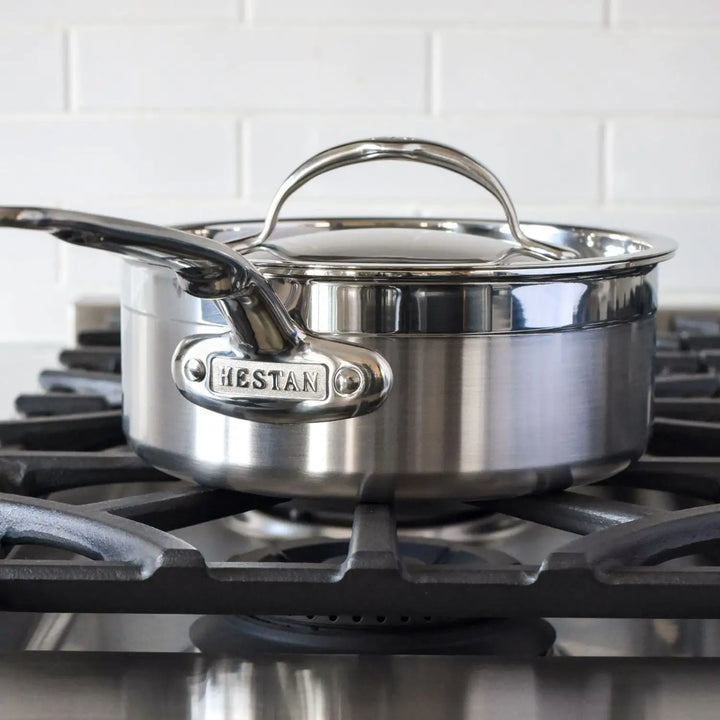 A ProBond Professional Clad Performance saucepan from the 28-piece set sits elegantly on a black stove grate, its tri-ply aluminum core ensuring even heat distribution. White subway tiles in the background add classic charm.