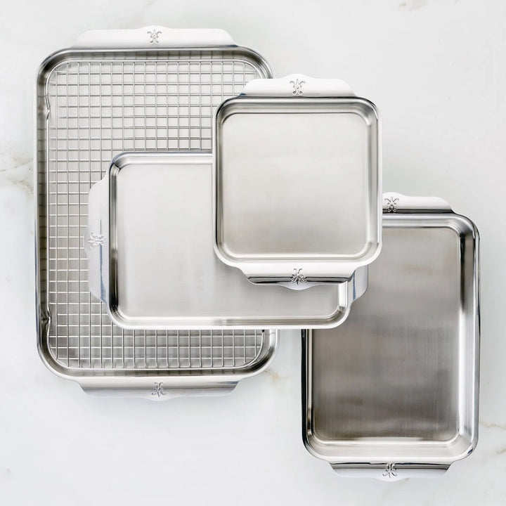 Four ProBond Professional Clad Performance baking sheets in stainless steel and one cooling rack are arranged on a white surface. The tri-ply aluminum core sheets vary in size, with one under the grid-patterned cooling rack.