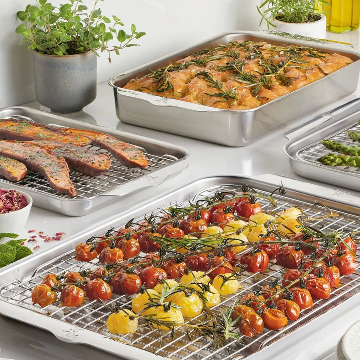 Several dishes are arranged on ProBond cooling racks with baking sheets. In the foreground, roasted cherry tomatoes and peppers, garnished with herbs, shine alongside cooked sausages, marinated chicken, and asparagus next to potted herbs—all made using the Professional Clad Performance 28-piece Set by ProBond.