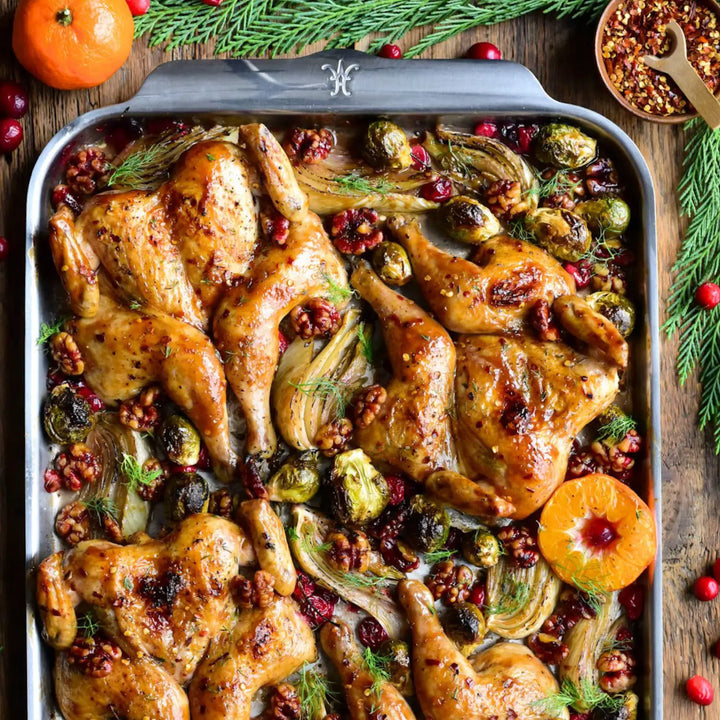 On a ProBond Professional Clad Performance baking sheet, a spatchcocked roasted chicken is surrounded by Brussels sprouts, fennel, cranberries, walnuts, and herbs. Nearby are a halved orange, a wooden spoon with red pepper flakes, and a tangerine on a rustic wooden table.