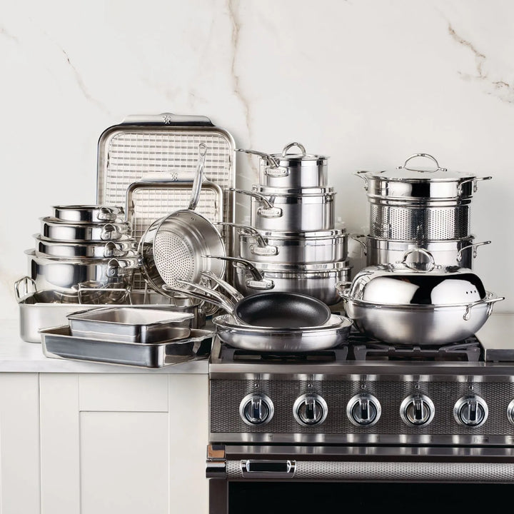 A 28-piece Professional Clad Performance stainless steel set by ProBond with tri-ply aluminum core is neatly arranged on the kitchen countertop. It includes pots, pans, baking sheets, and a strainer in front of a modern stovetop with multiple dials.