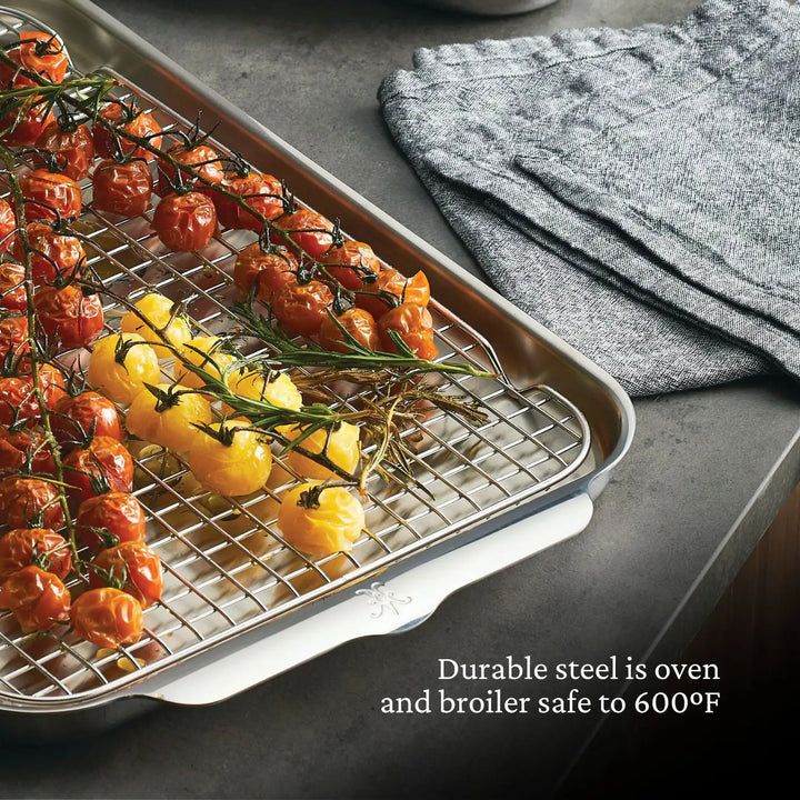 On a gray countertop, roasted red and yellow tomatoes with rosemary sit on a baking sheet with a cooling rack, near folded gray cloths, featuring durable ProBonds Professional Clad Performance 28-piece Set stainless steel cookware, oven and broiler safe up to 600°F.