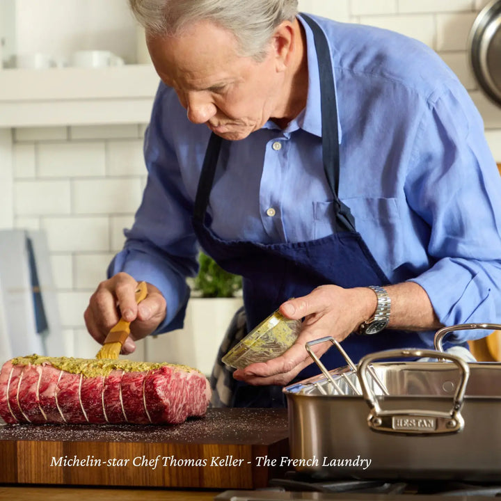 A person in a blue shirt and apron brushes marinade onto raw meat on a wooden counter. A pot from the ProBond Professional Clad Performance 28-piece Set is nearby, with white tiled walls and kitchen items in the background enhancing the professional vibe with stainless steel cookware.