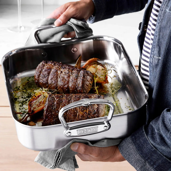 A person in a denim jacket and striped shirt holds a ProBond Professional Clad roasting pan (from the 28-piece set) with a tri-ply aluminum core, containing two beef roasts garnished with herbs and roasted onions, while a gray cloth drapes over its handle.