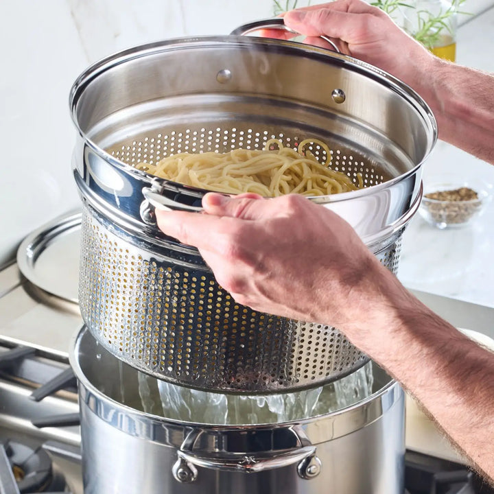 A person expertly drains spaghetti using a ProBond pot with a built-in strainer from the Professional Clad Performance 28-piece Set over the stove, steam swirling as the gas burner glows beneath. A plant and small bowl add charm to the background.