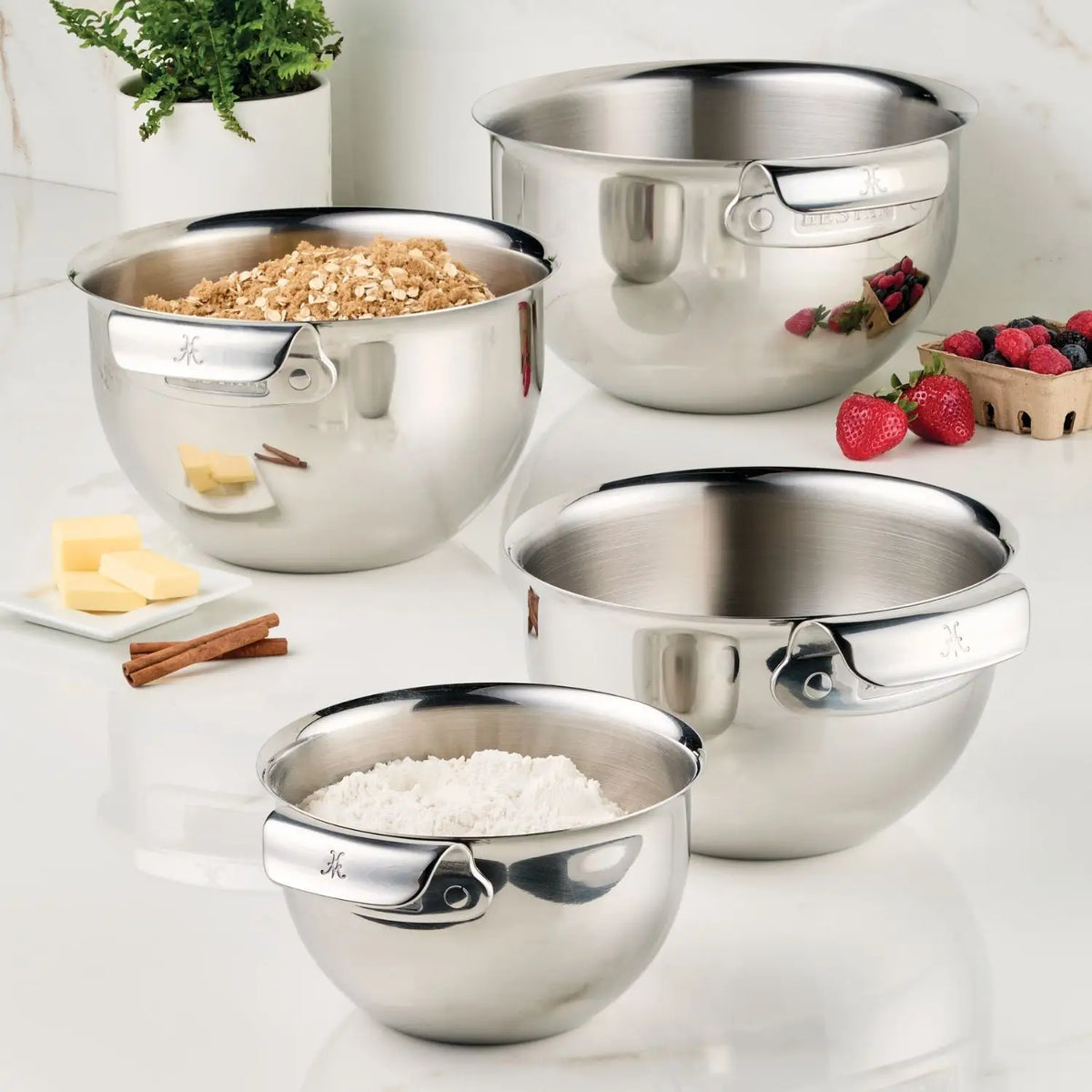 Four mixing bowls from the NanoBond Titanium x Tyler Florence 41-Piece Cookware Set are arranged on a white countertop, with two holding flour and oats. Nearby sit fresh strawberries, butter, cinnamon sticks, and a small basket of berries.