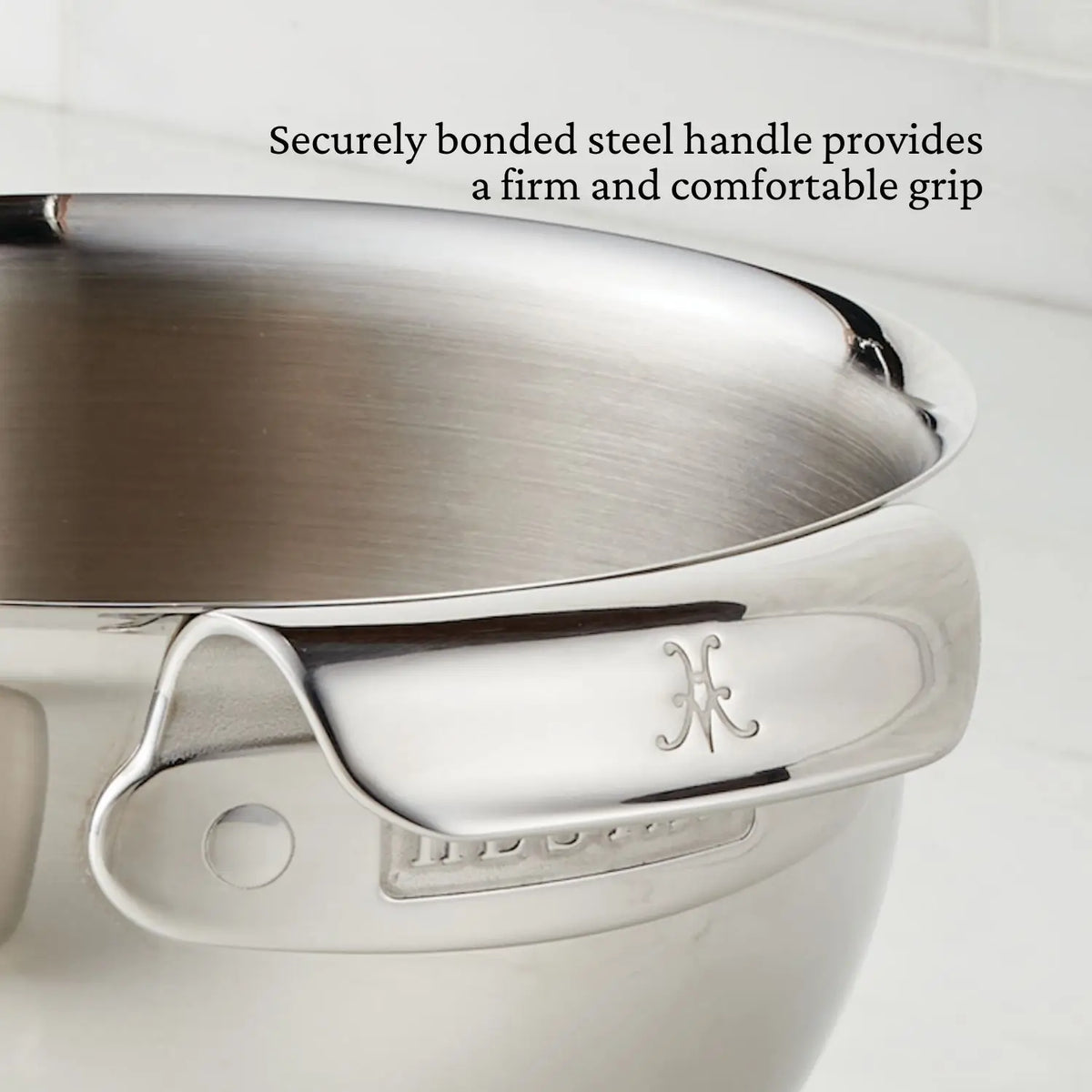 A close-up of a stainless steel mixing bowl from the NanoBond Titanium x Tyler Florence Cookware Set, 41-Piece, with a securely bonded steel handle and engraved logo. Text above: “Securely bonded steel handle provides a firm and comfortable grip.”.