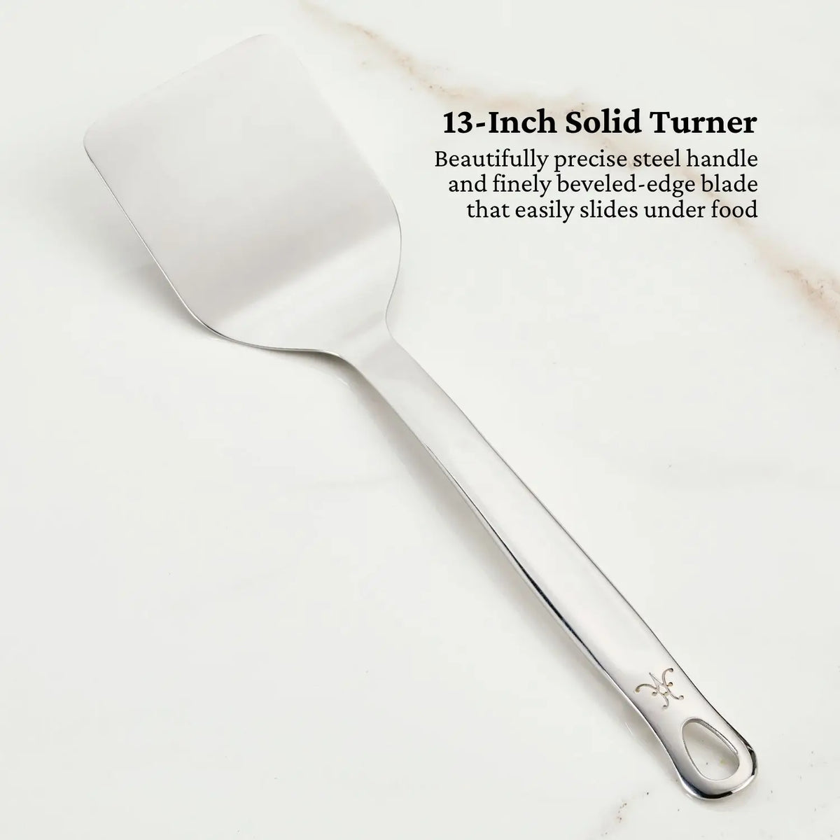 A tri-ply stainless steel 13-inch solid turner, part of the OvenBond Epicurean 21-Piece Set by Hestan Provisions, lays on a white surface. The handle features a small decorative emblem. Text reads: Beautifully precise steel handle and finely beveled-edge blade that effortlessly slides under food.