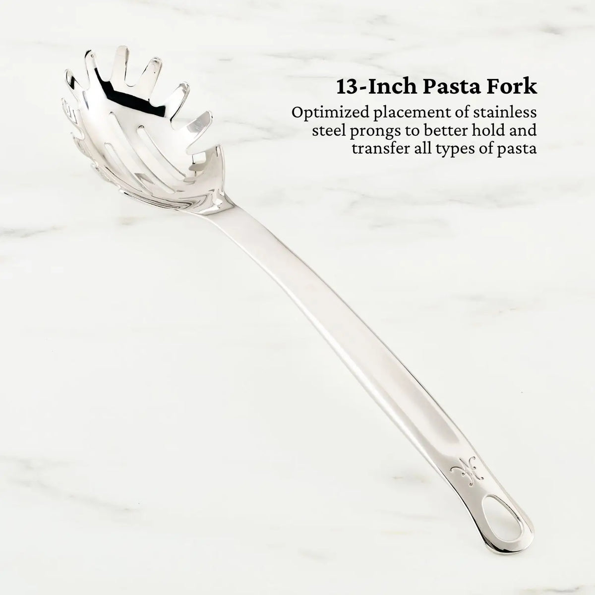 A Hestan Provisions pasta fork with pronged edges, designed for effortless pasta handling, sits on a light marble surface. The inscription beside it reads: 13-Inch Pasta Fork in tri-ply stainless steel. Its part of the OvenBond Epicurean 21-Piece Set.