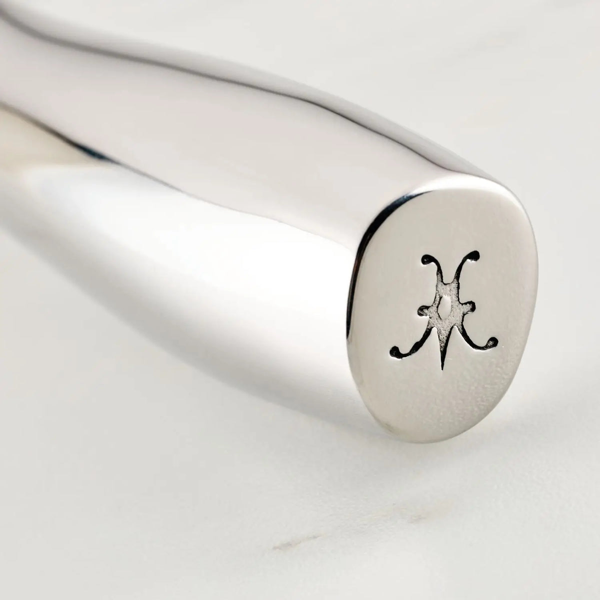The end of a Hestan Provisions OvenBond Tri-ply Epicurean stainless steel knife handle on a white surface features an engraved decorative symbol akin to a flower or star, showcasing its polished, premium craftsmanship.