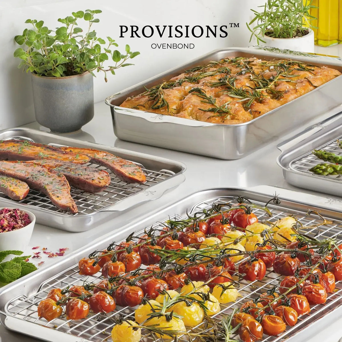 An assortment of food is on the countertop. Roasted cherry tomatoes and rosemary are on Hestan Provisions OvenBond Tri-ply Epicurean Stainless Steel bakeware, with marinated meat nearby. A dish of baked chicken, a small plant, and a bottle of oil complete the inviting scene.