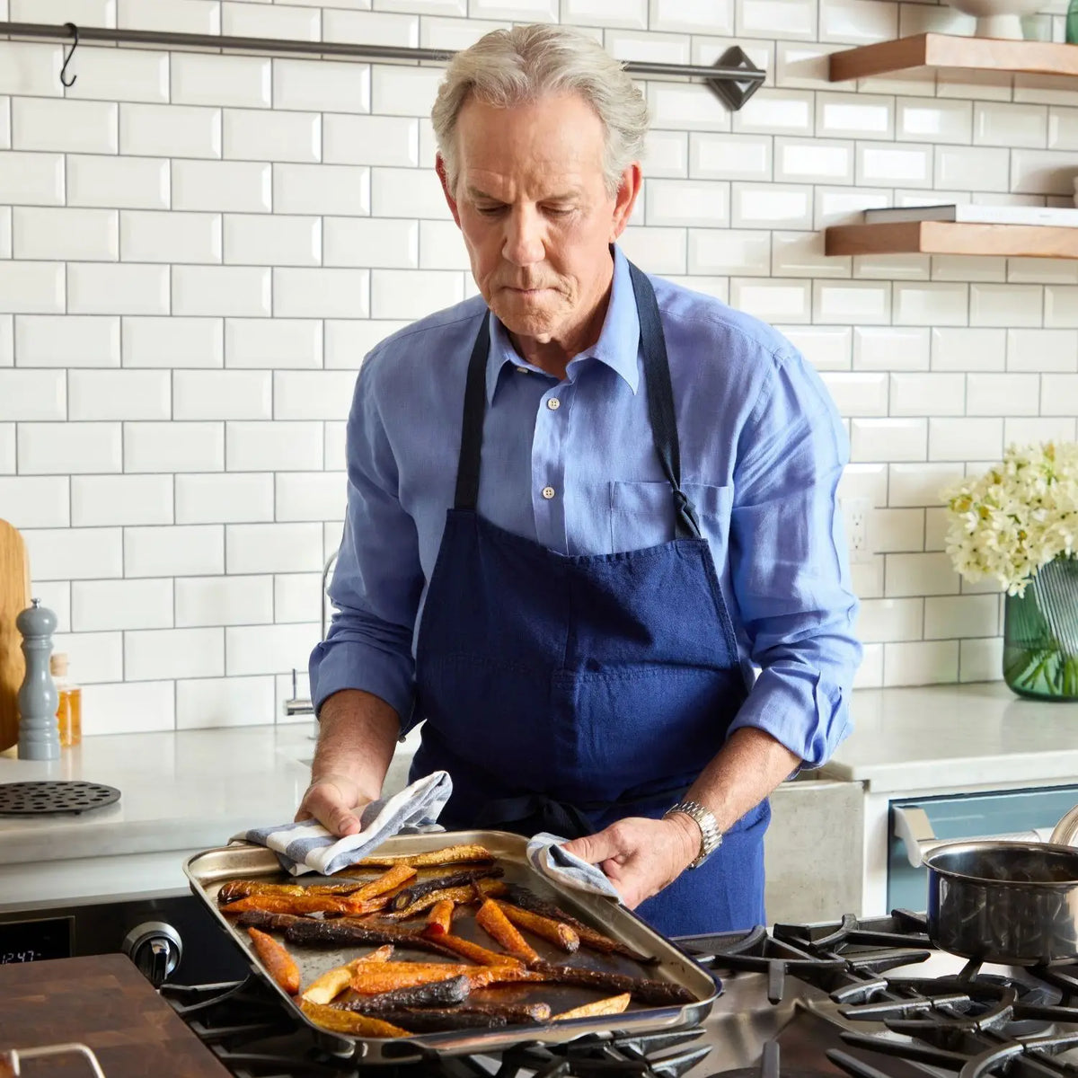 An older man with gray hair, in a blue shirt and apron, proudly presents roasted veggies in his modern kitchen with white subway tiles, shelves, and flowers; he relies on the Hestan Provisions OvenBond Tri-ply Epicurean Stainless Steel 21-Piece Set for perfect results.
