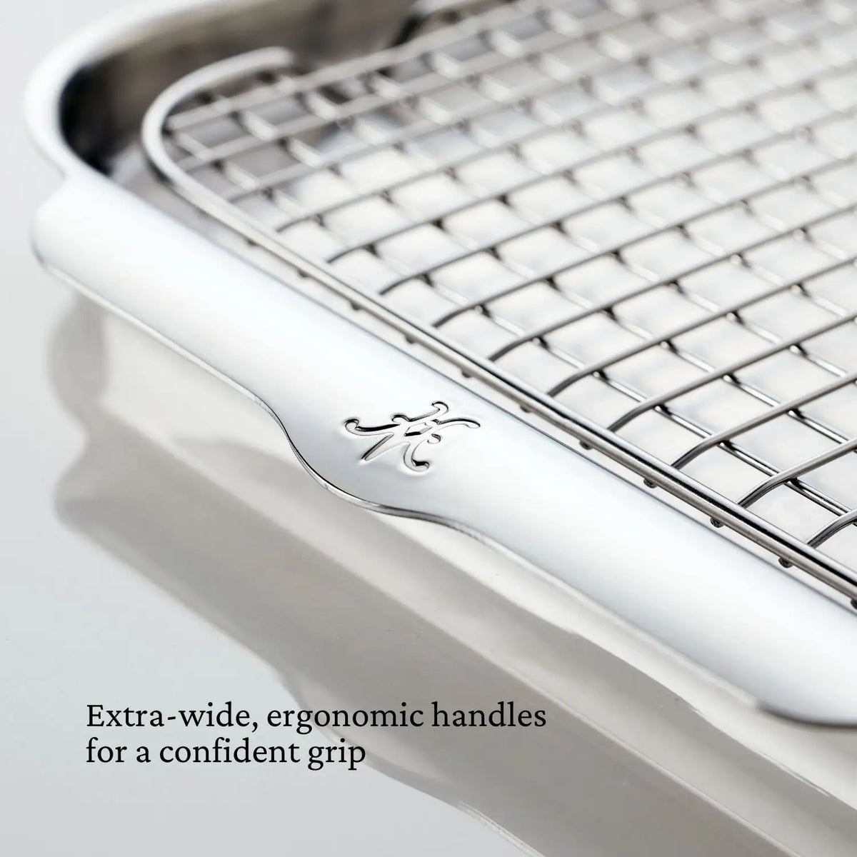 Close-up of the rectangular metal cooling rack from the Hestan Provisions OvenBond Tri-ply Epicurean Stainless Steel 21-Piece Set, showcasing extra-wide, ergonomic handles with an engraved logo. Text reads: Extra-wide, ergonomic handles for a confident grip. It features a grid pattern in durable tri-ply stainless steel.