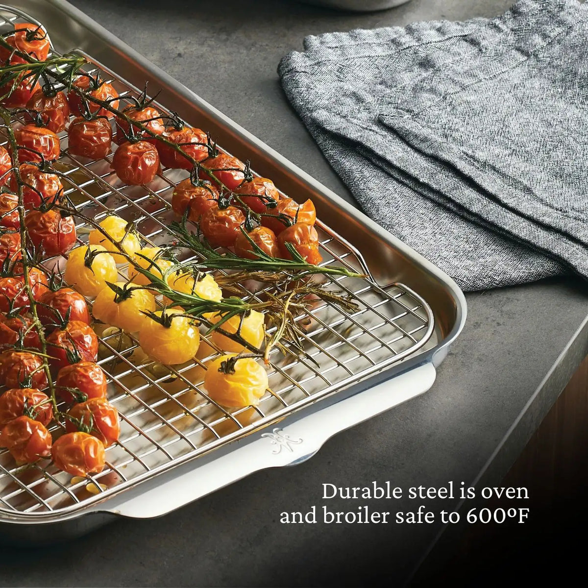 A stainless steel roasting pan from the OvenBond Tri-ply Epicurean Stainless Steel 21-Piece Set by Hestan Provisions cradles vine cherry tomatoes. It sits on a gray countertop beside folded fabric. The text highlights its durability, being safe up to 600°F in ovens and under broilers.