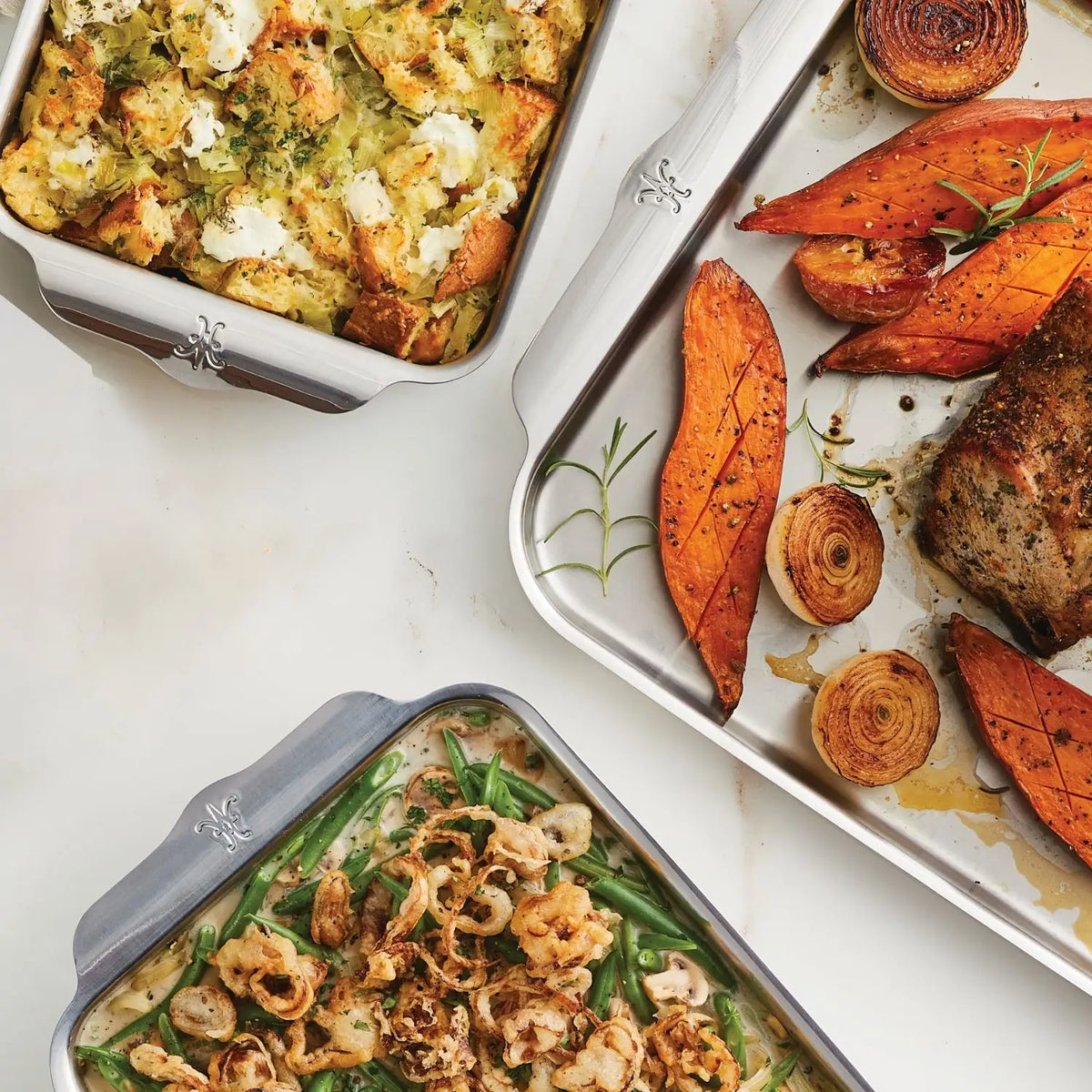 Resting on a light marble surface are three dishes: herb-topped stuffing with cheese in durable bakeware, roasted sweet potatoes and onions with meat in a tray, and green bean casserole with crispy onions, all crafted using the Hestan Provisions OvenBond Tri-ply Epicurean Stainless Steel 21-Piece Set.