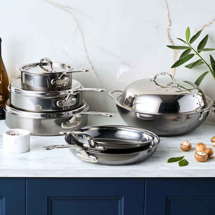 A collection of the 10-piece Limited Edition Winter Titanium Cookware Set by NanoBond, featuring sleek handles, is artfully displayed on a marble surface. Nearby are a few mushrooms, a small green plant, and a wine bottle. The scene includes a white marble wall and a blue cabinet adding color below.