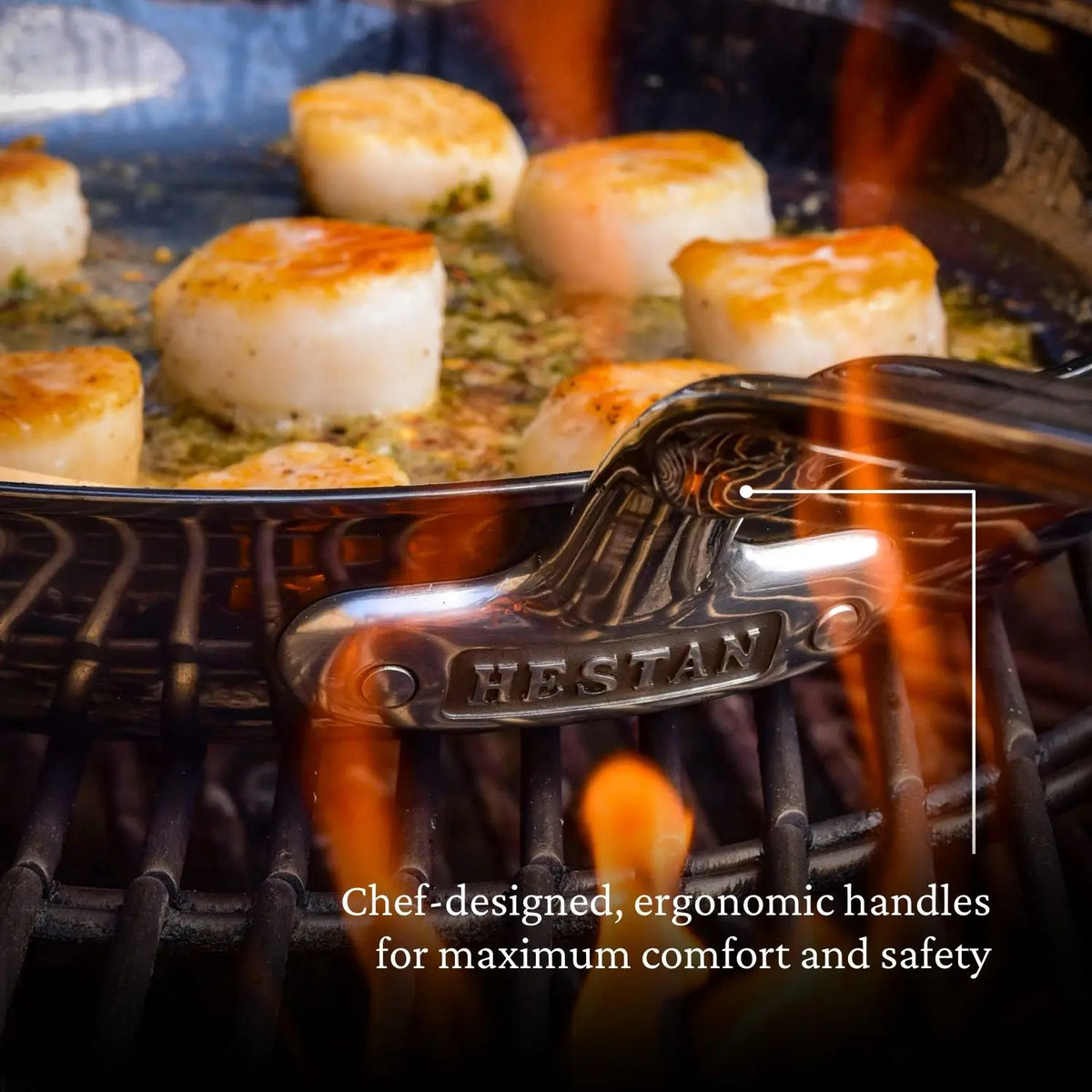 A NanoBond Titanium Skillet, engraved with “HESTAN,” sits on a grill cooking scallops in oil as orange flames flare. Text overlay: “Chef-designed, ergonomic handles for maximum comfort and safety.”.