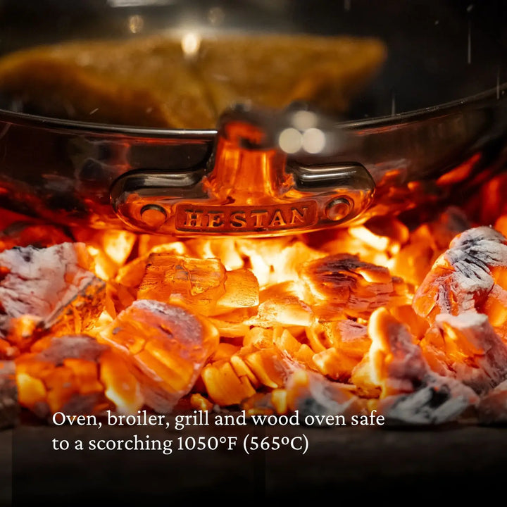 Close-up of glowing hot coals under a metal pan, with HESTAN on the handle, reveals the Titanium Skillets by NanoBond. Made from Molecular Titanium, this skillet is oven, broiler, grill, and wood oven safe up to 1050°F (565°C).