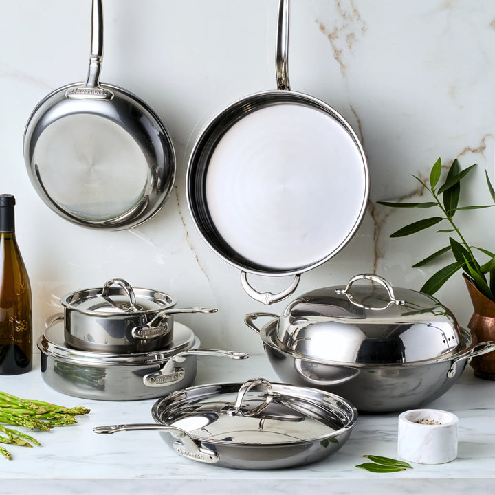 The 10-piece NanoBond Limited Edition Winter Titanium Cookware Set, including stainless steel frying pans, saucepans with lids, and a large pot, is displayed on a marble countertop. Some pots are stacked and two pans hang elegantly on the wall with a wine bottle and herbs adding charm to the scene.