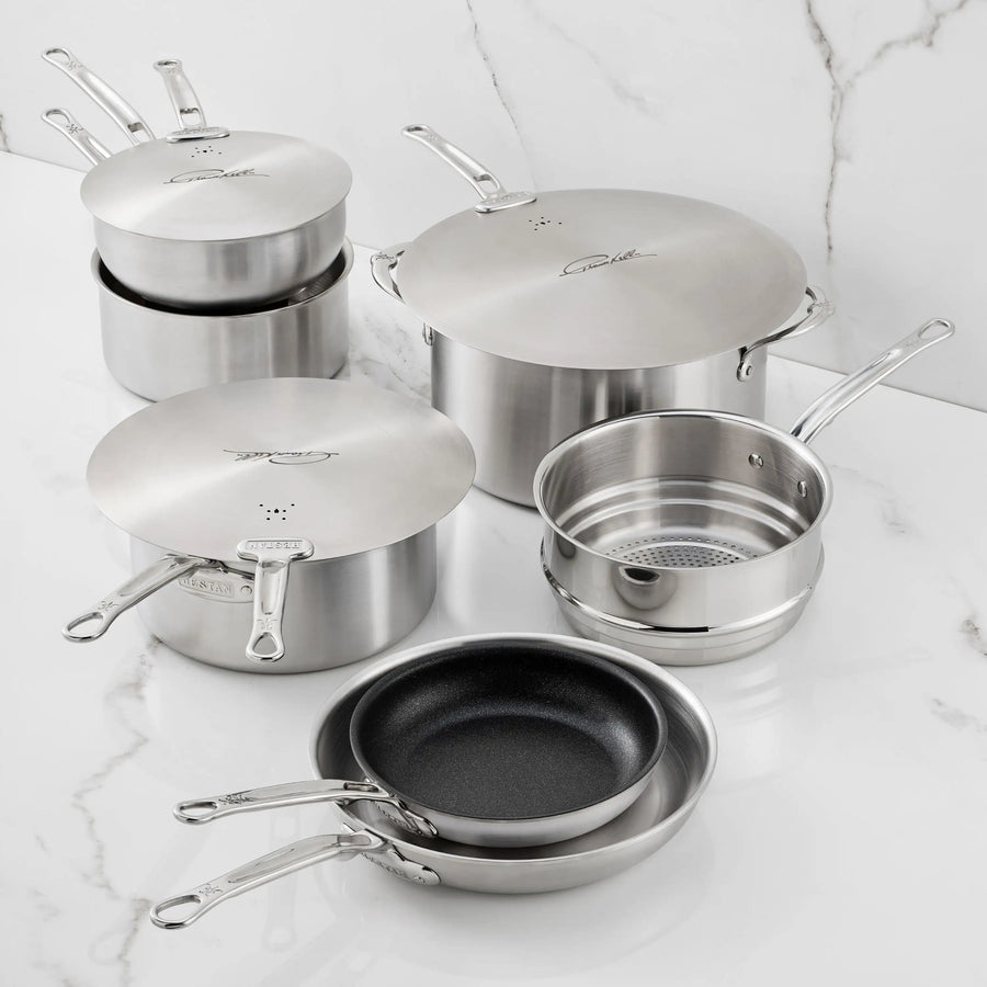 The Thomas Keller Commercial Clad Stainless Steel 10-Piece Cookware Set, including pots with lids, a saucepan with steamer insert, and two frying pans (one nonstick), is elegantly displayed on a white marble countertop.