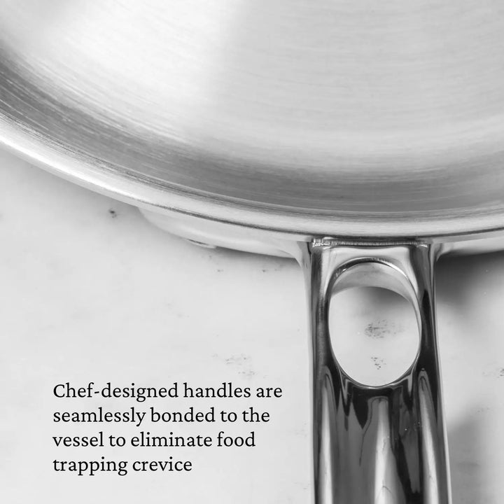 Close-up of the Commercial Clad Stainless Steel pan handle shows seamless bonding. Text overlay: Chef-designed handles are seamlessly bonded to the Tri-Ply Stainless Steel vessel to eliminate food-trapping crevices, inspired by Thomas Keller Insignia Cookware.