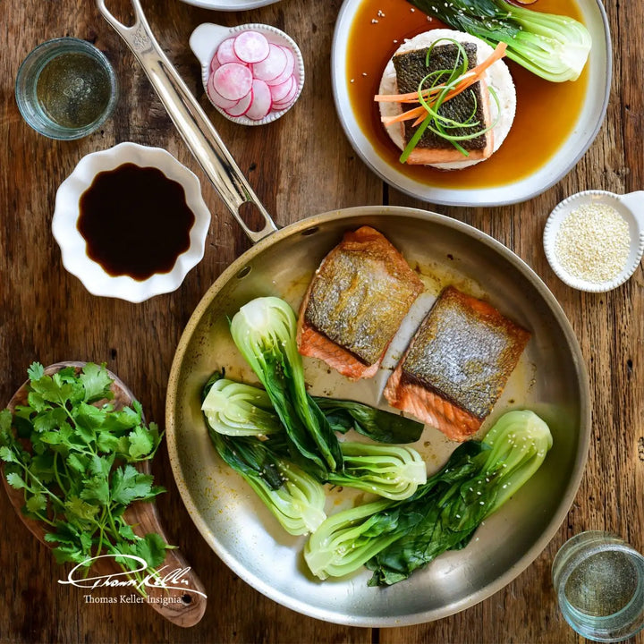 A wooden table presents a plate of salmon fillets and bok choy, expertly made using the Thomas Keller Insignia Commercial Clad Stainless Steel 10-Piece Cookware Set. Nearby, soy sauce and radishes complement the dish with cilantro sprigs, and a glass of water is partially visible.