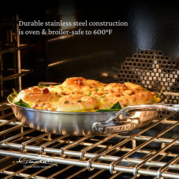 A Thomas Keller Insignia stainless steel skillet with chicken and vegetables is in the oven. Text above reads, Durable Tri-Ply Stainless Steel Cookware construction is oven & broiler-safe to 600°F. The Commercial Clad Signature logo is visible in the bottom left.