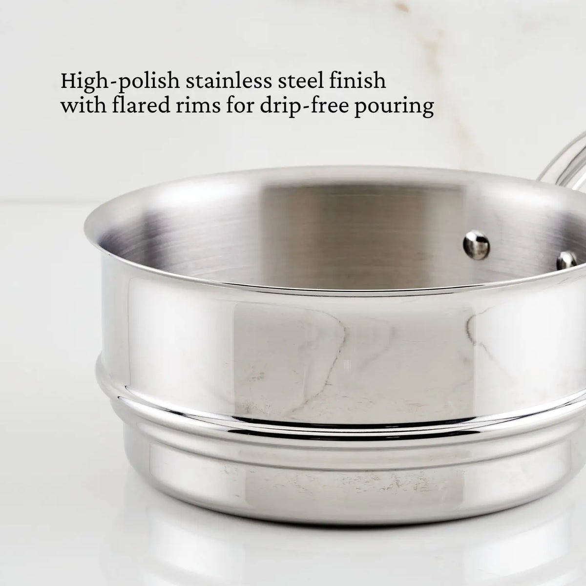 A Thomas Keller Commercial Clad Stainless Steel 10-Piece Cookware Set sits on a white surface, featuring flared rims for drip-free pouring and a high-polish finish that reflects its durable, professional craftsmanship.