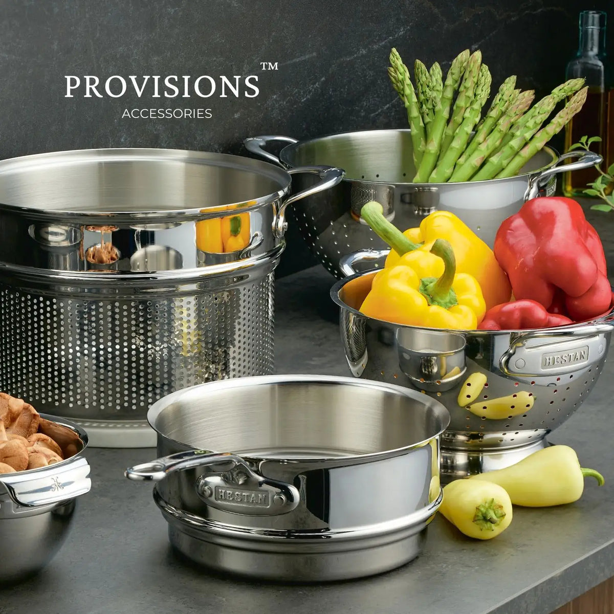 A Thomas Keller Commercial Clad Stainless Steel 10-Piece Cookware Set sits on the countertop, filled with asparagus, peppers, and mushrooms. "PROVISIONS" appears in the background, inspiring a professional kitchen atmosphere.