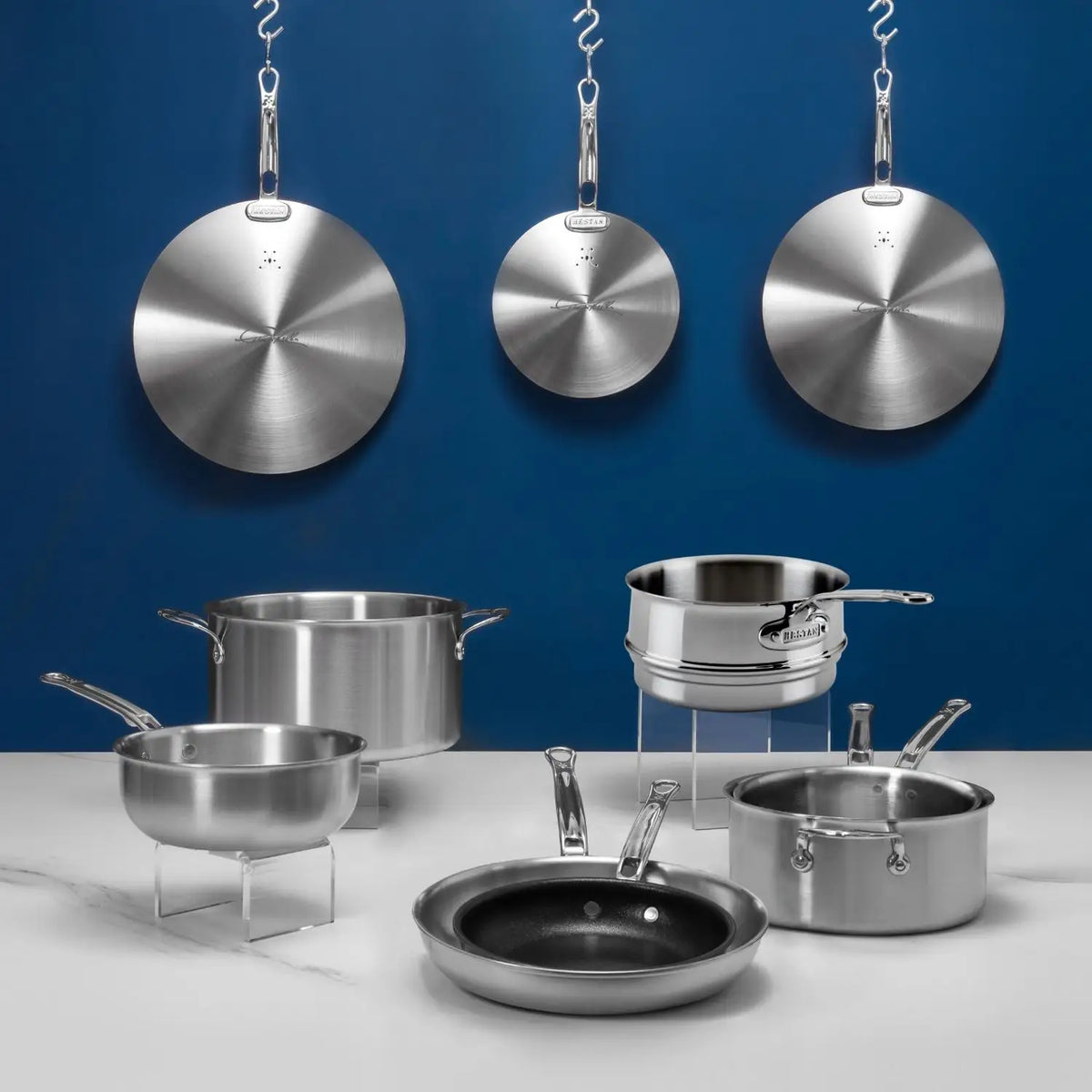 The Thomas Keller Commercial Clad Stainless Steel 10-Piece Cookware Set, featuring pots, pans, and lids, is arranged on a white surface with three lids hanging on a blue wall in the background.