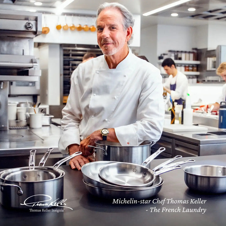 A chef in a white coat stands in a professional kitchen, surrounded by the Thomas Keller Insignia Commercial Clad Stainless Steel 10-Piece Cookware Set. Several people work busily in the background, highlighting Michelin-star chef Thomas Keller and his cookware preference.
