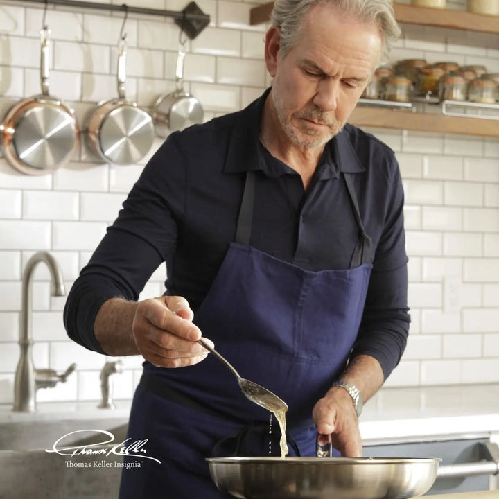 A man in a navy apron cooks using the Thomas Keller Insignia Commercial Clad Stainless Steel 10-Piece Cookware Set, gently pouring liquid from a spoon into a pan. The kitchen has white tile walls and copper pans, with the Thomas Keller Insignia visible in the lower left corner.
