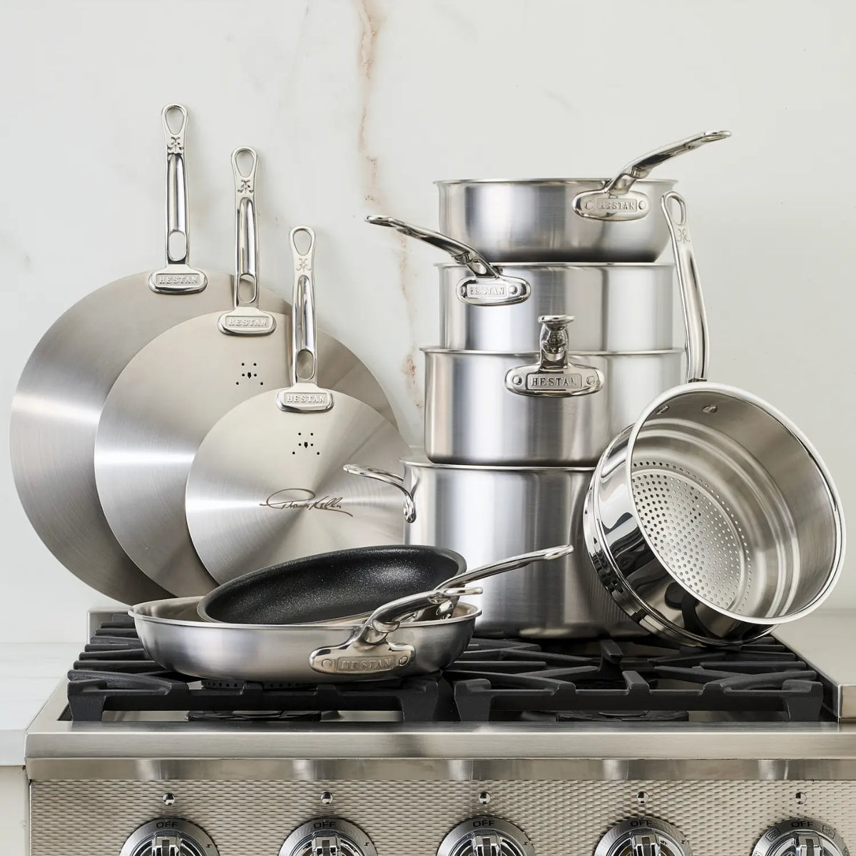 The Thomas Keller Commercial Clad Stainless Steel 10-Piece Cookware Set, featuring pots, pans, lids, and a strainer, is elegantly stacked and displayed on a stovetop against a light kitchen backsplash.