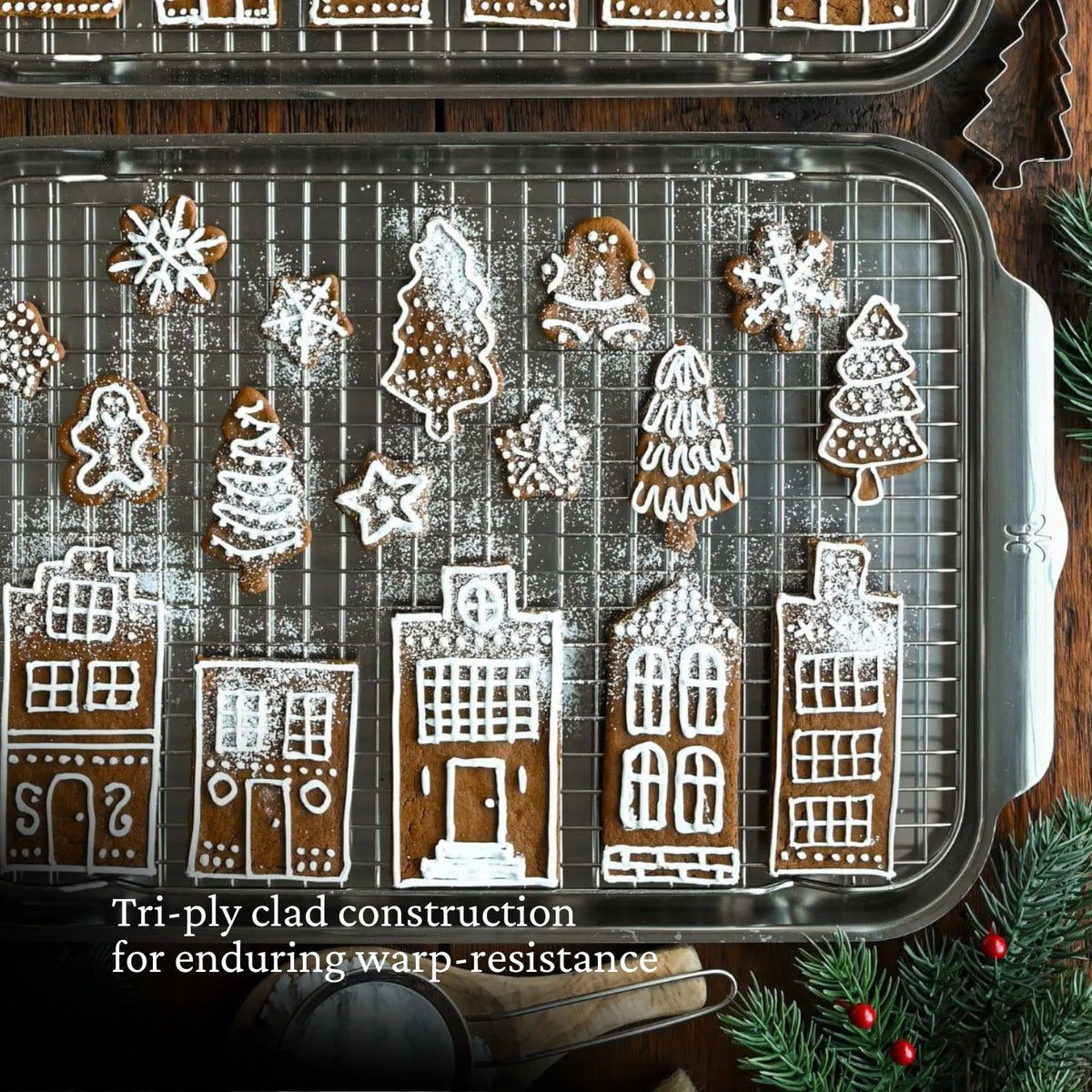 A durable bakeware from Hestan Provisions features gingerbread cookies shaped like houses, trees, stars, and bells with white icing. On a cooling rack with pine and red berries beneath it rests the OvenBond Tri-ply Epicurean Stainless Steel 21-Piece Set.