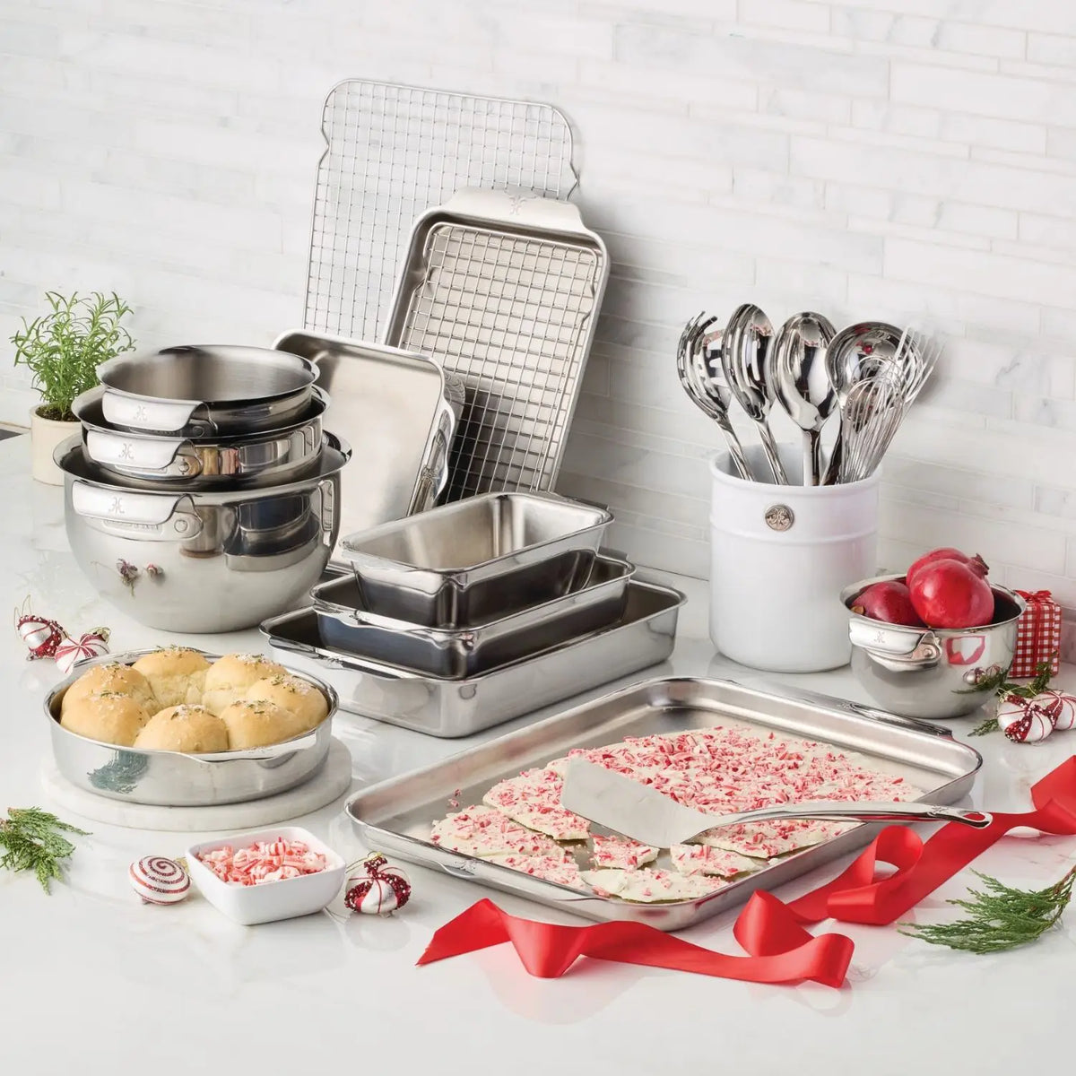 A kitchen countertop displays a Hestan Provisions OvenBond Tri-ply Epicurean Stainless Steel 21-Piece Set, baking sheets, mixing bowls, and serving utensils. Buns, peppermint bark, candies adorn the area with a red ribbon and rosemary sprig, showcasing the charm of quality bakeware.
